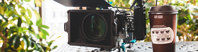 A professional film camera with a microphone attachment sits next to a coffee cup on an outdoor table surrounded by foliage