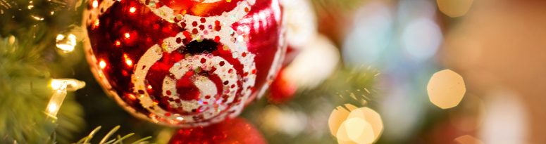 A red Christmas bauble decorated with white and glittery swirl patterns, hanging from a decorated fir tree branch