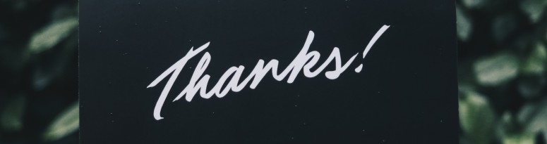 a black card with the word 'Thanks!' written in white cursive font to show gratitude, set against a background of lush green foliage.